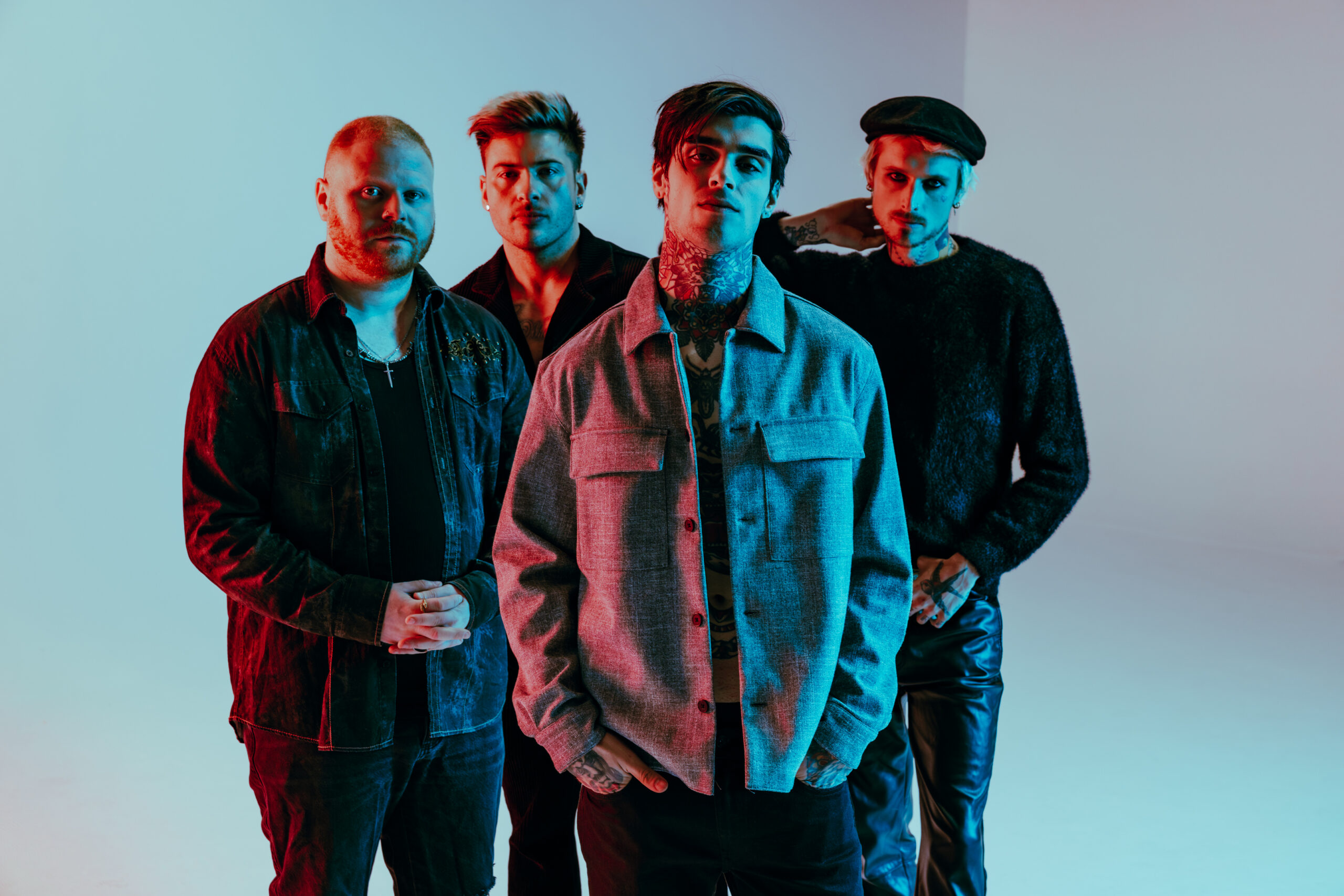 Four men with tattoos and stylish outfits stand together under colorful lighting