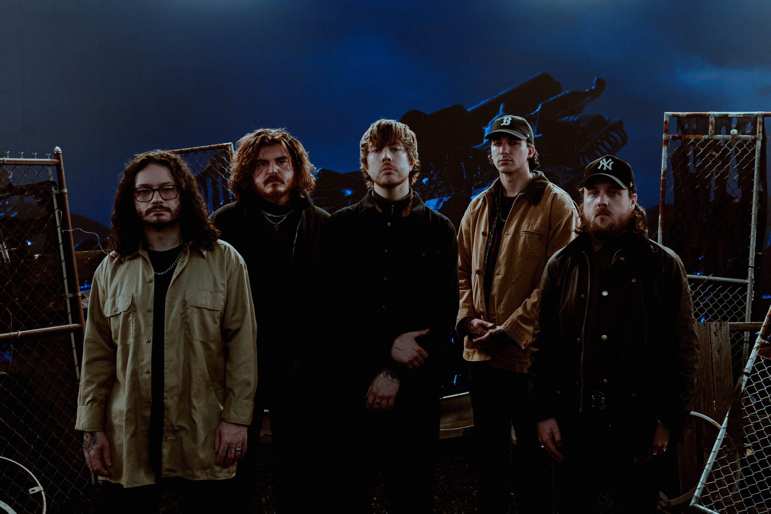 Five men in casual wear stand in front of a dark, industrial background.